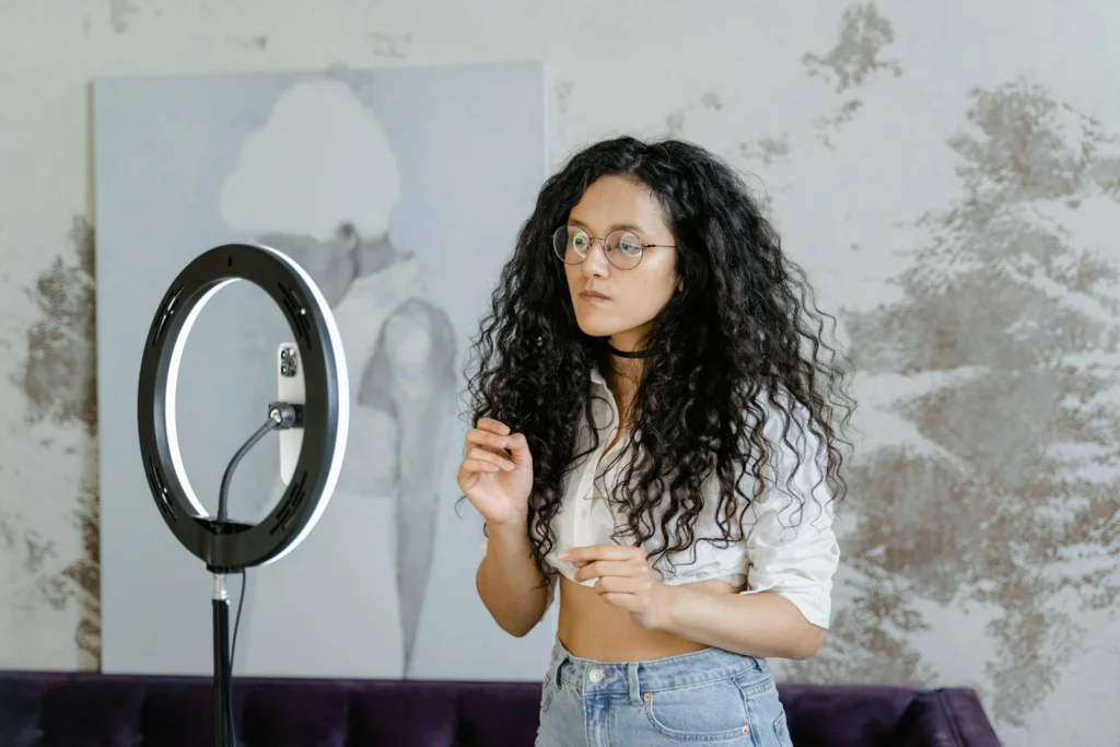 A young woman stands indoors, using a ring light and smartphone for streaming content creation.