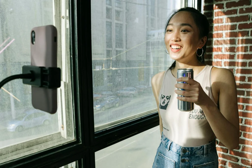 Young Asian woman smiling during a livestream, holding a soda can indoors near a bright window.