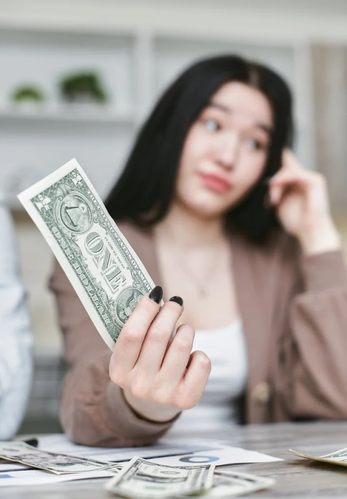 Young woman with dark hair holding a one-dollar bill, seated at a table.