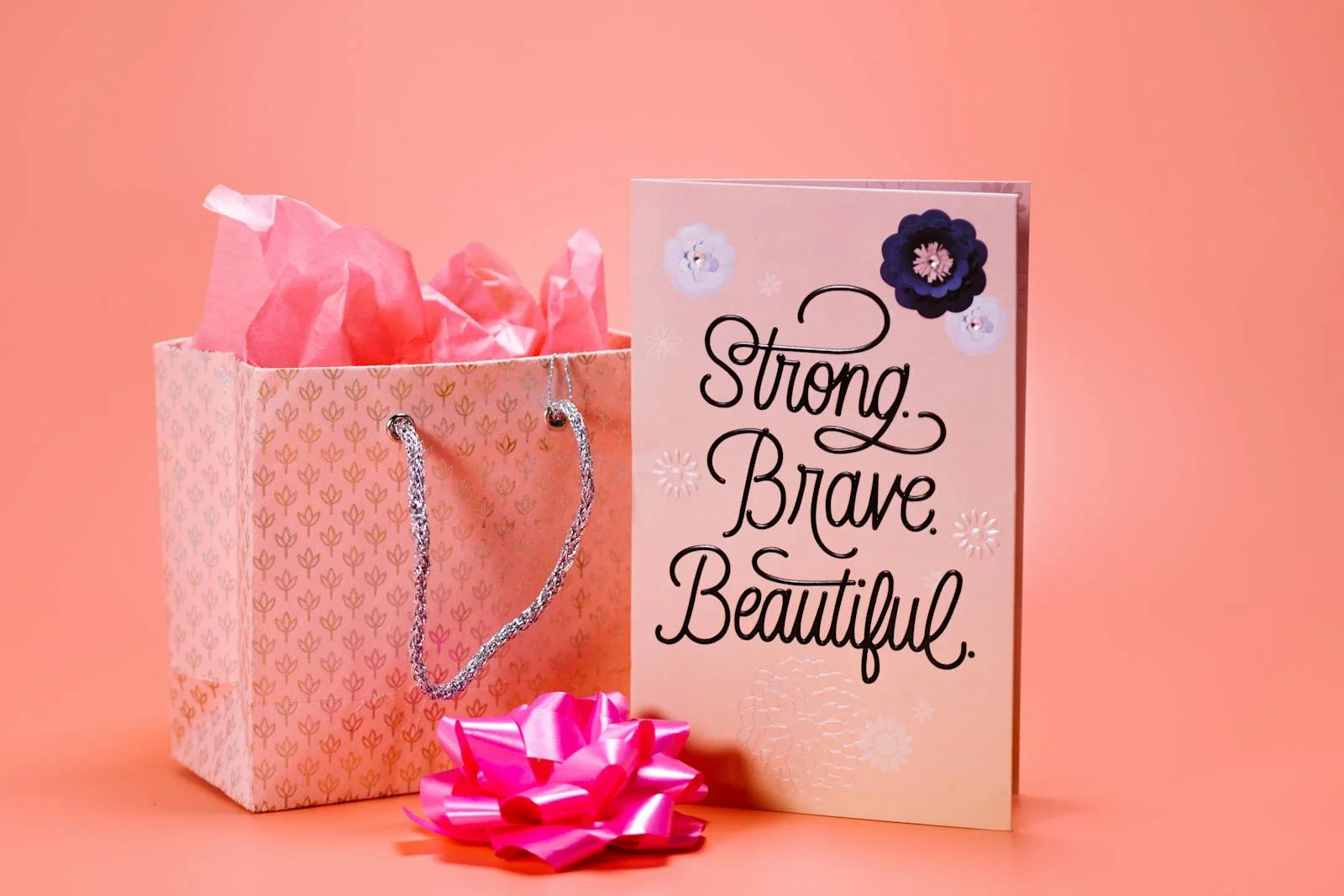 Pink gift bag and card with 'Strong. Brave. Beautiful.' text on a peach background.