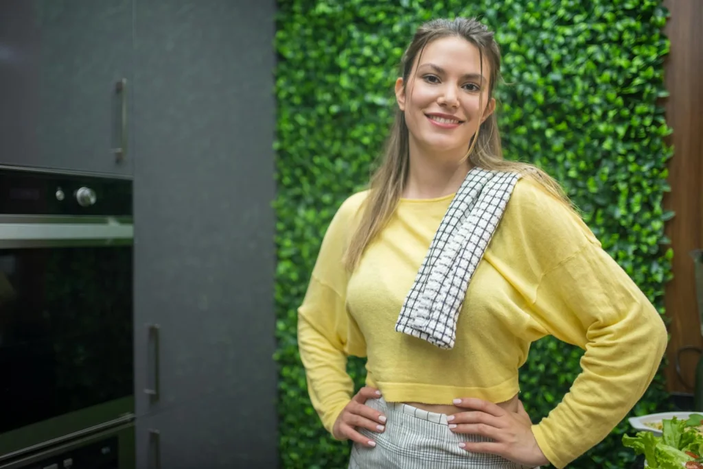 Smiling woman in a yellow sweater posing in a modern Portuguese kitchen with lush greenery backdrop.
