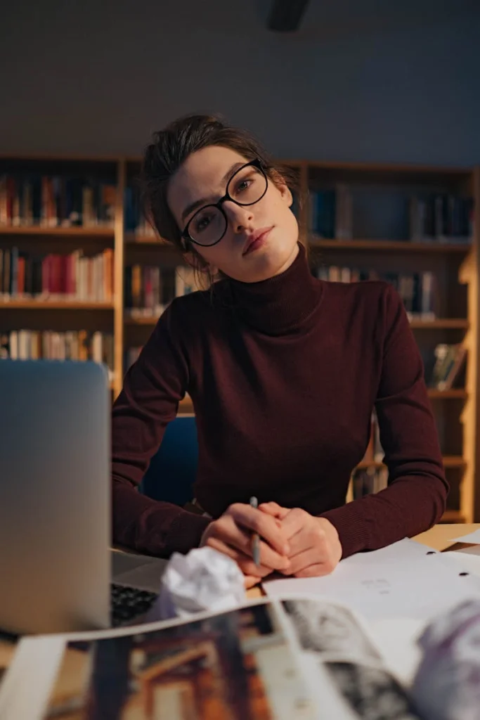 A woman in glasses studies intently at a library work desk, surrounded by books.