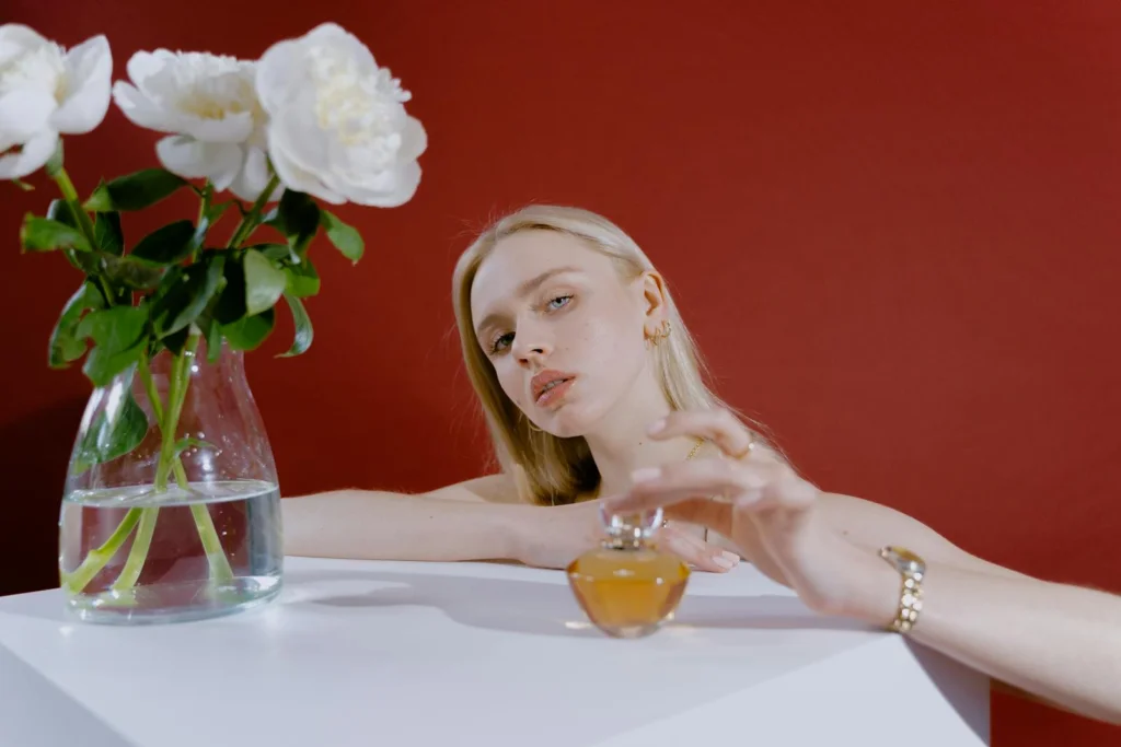 A woman with blond hair and a perfume bottle next to a vase of white roses on a white table.