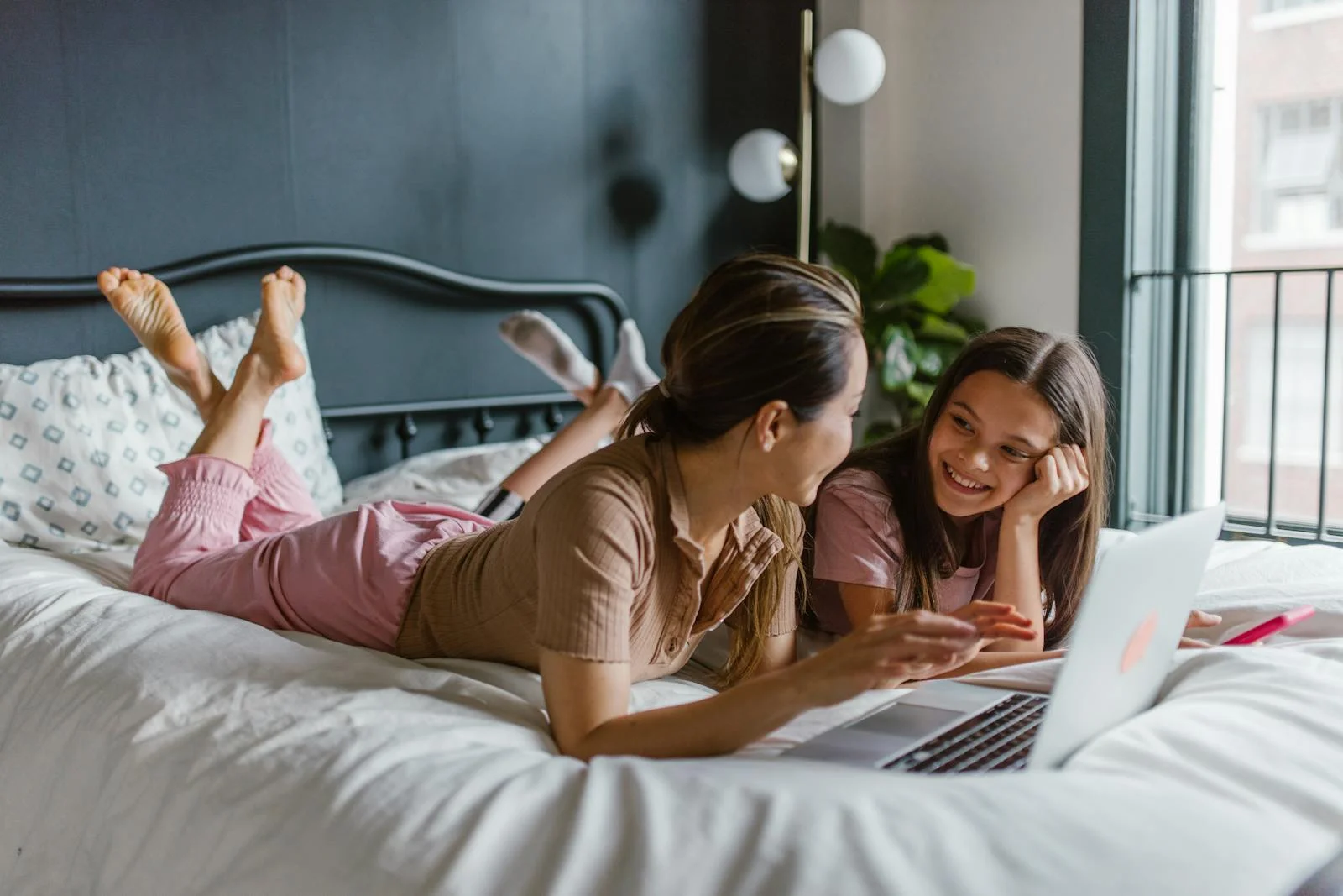 A mother and daughter sharing a joyful moment together, lying on a bed with a laptop.