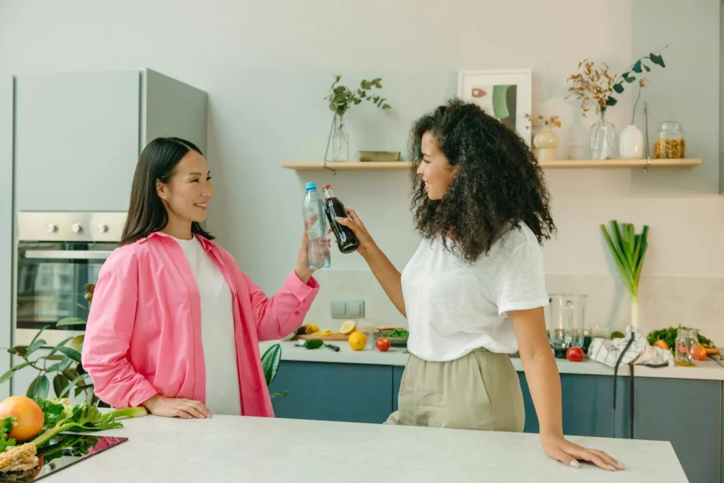 Two women in a kitchen clinking a soda bottle and water bottle, enjoying a healthy lifestyle moment.