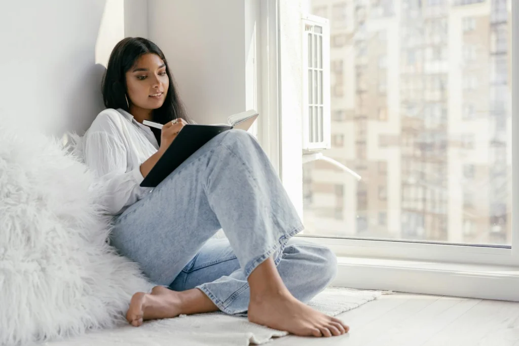 Young woman wearing denim and white, reading and writing by the window, enjoying a peaceful afternoon indoors.