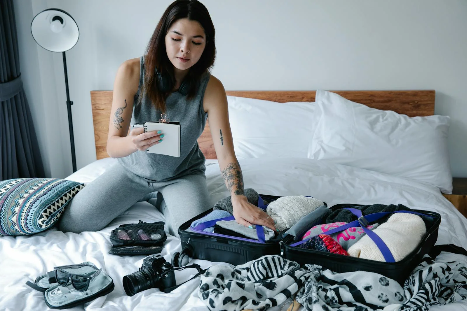Woman sitting on bed packing a suitcase and taking notes for her travel plan.
