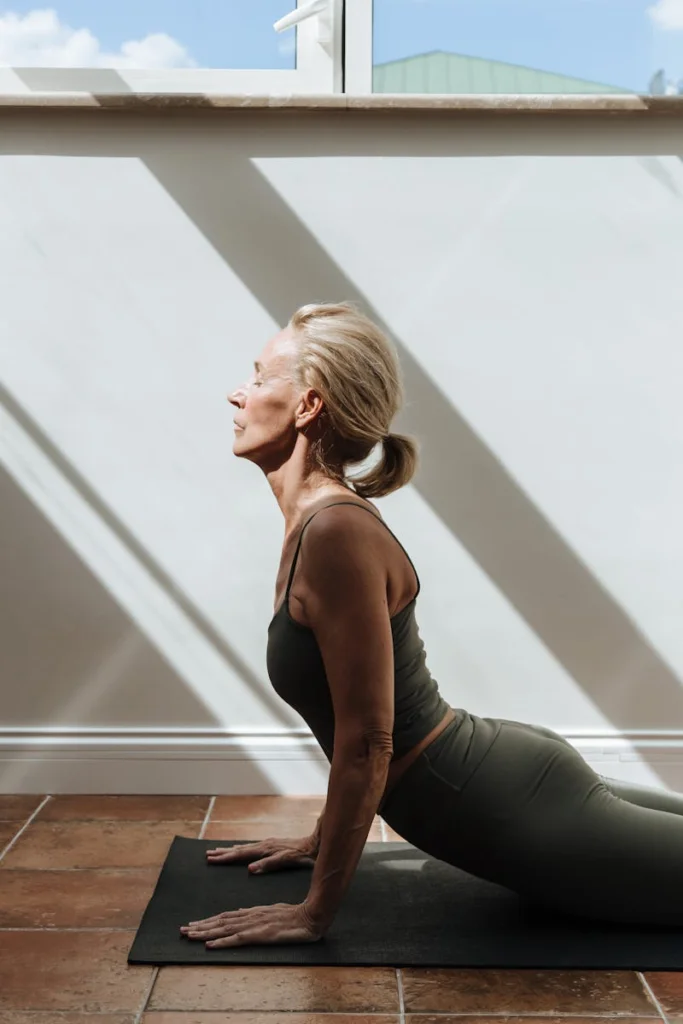 Elderly woman practicing yoga indoors with sunlight streaming through windows, embodying health and fitness.