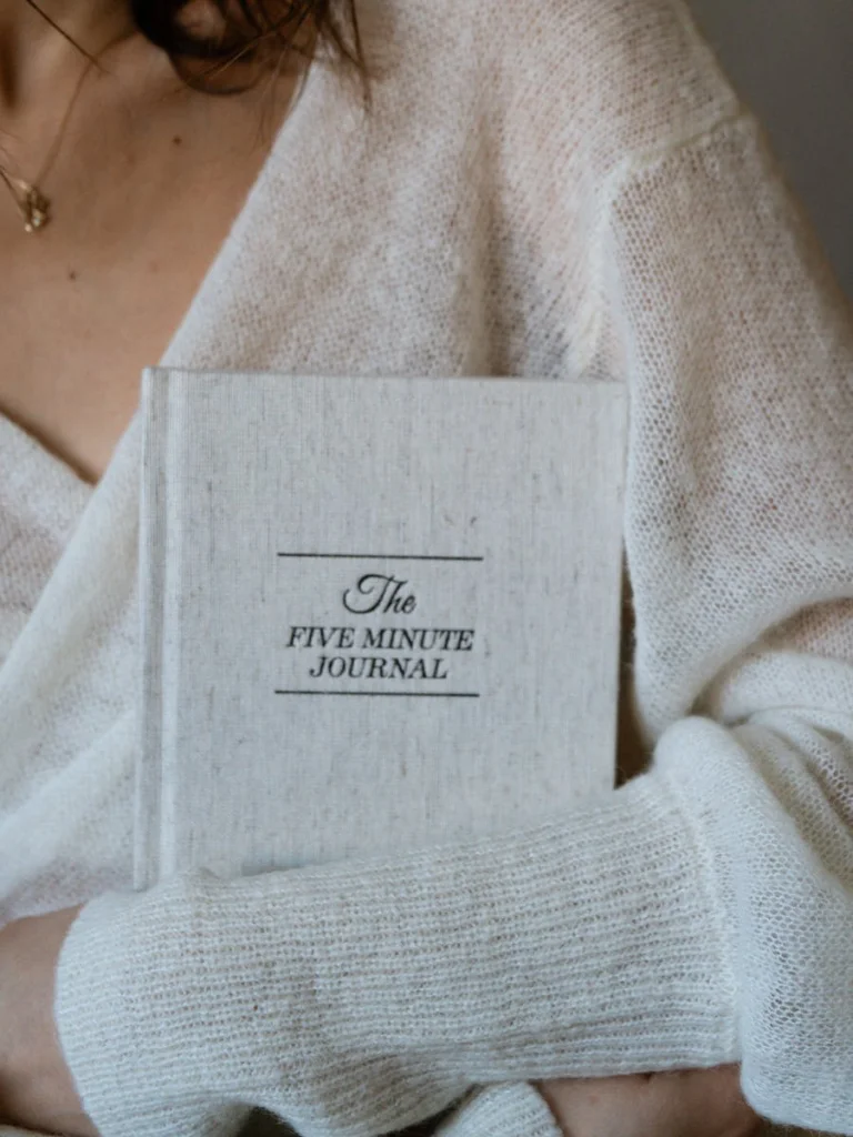 Close-up of a woman in a sweater holding The Five Minute Journal.