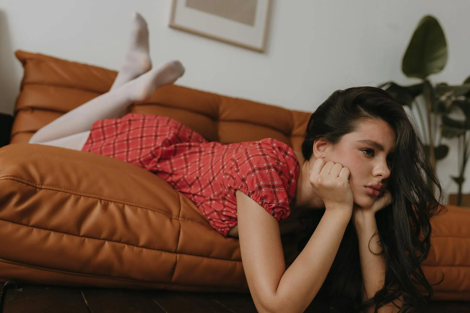Brunette woman in red dress lying on a brown sofa indoors, looking thoughtful.