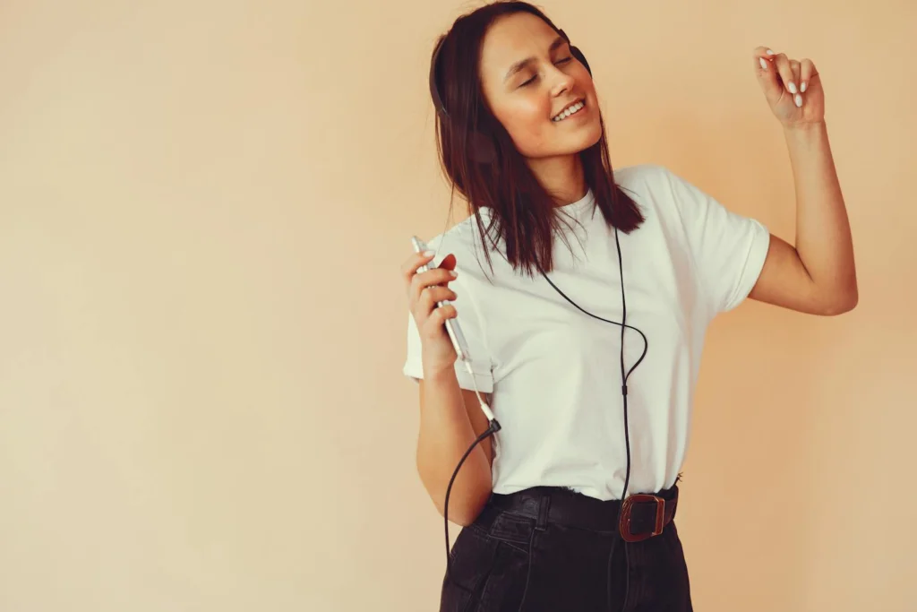Happy young female in casual clothes smiling and dancing with eyes closed while listening to good music in headphones in studio on beige background