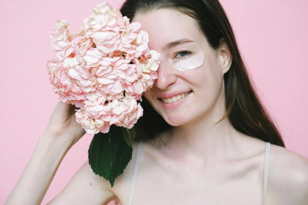 Cheerful woman with skincare cream and hydrangea against pink background, conveying natural beauty.