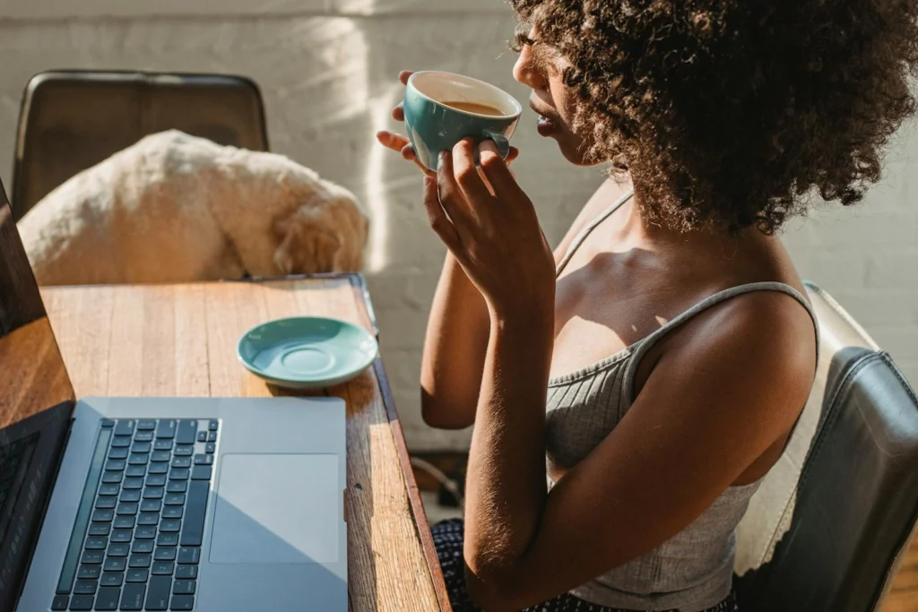 A woman enjoys coffee while working on a laptop at home with her dog nearby.