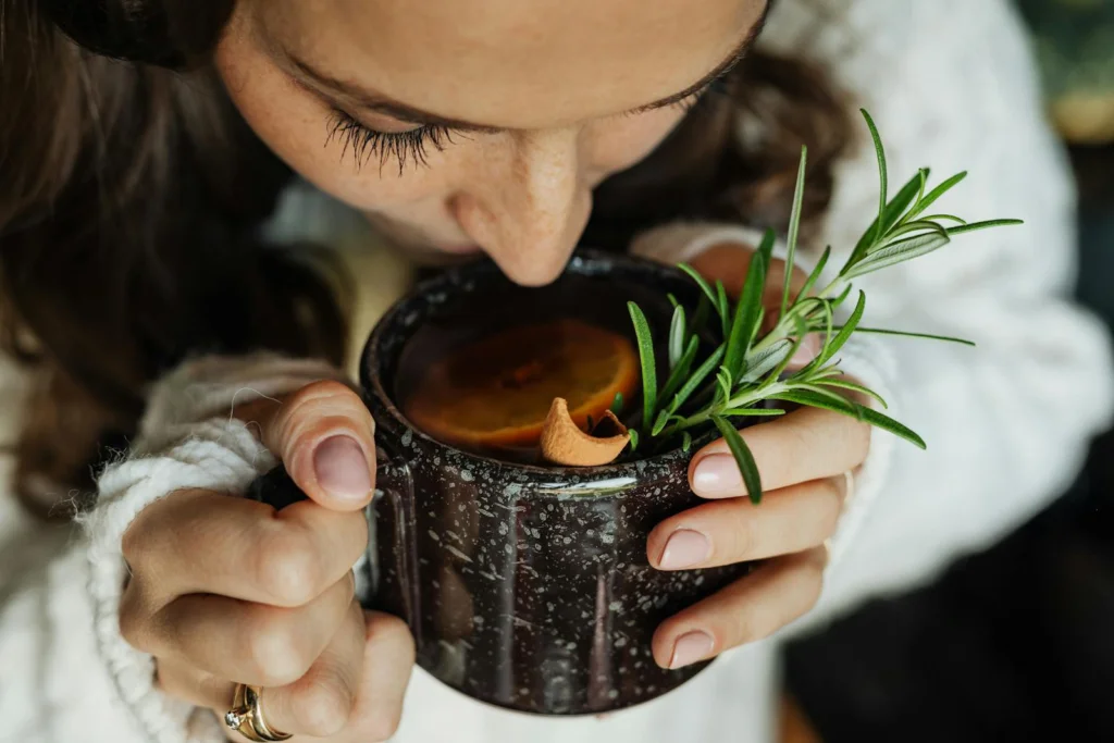 Woman savoring hot herbal tea with rosemary and orange, close-up view.