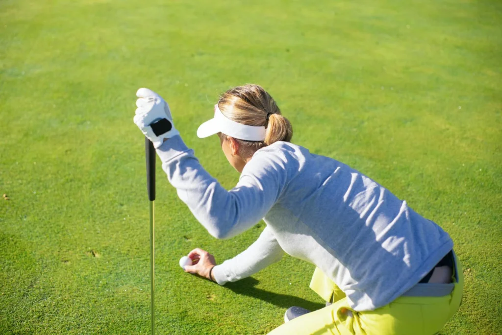 Female golfer in a gray sweater prepares to putt on a bright day outdoors.