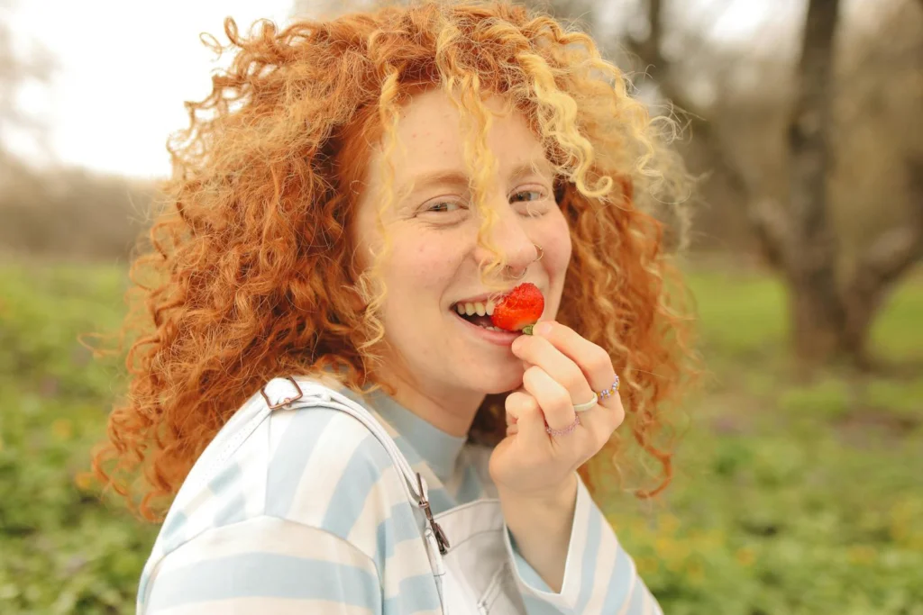 Smiling woman with curly red hair eating a strawberry in a sunny outdoor setting.
