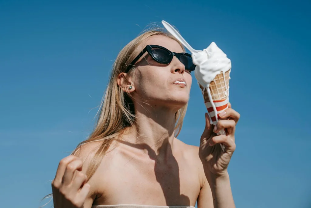 A woman wearing sunglasses enjoys a melting ice cream cone against a clear blue sky.