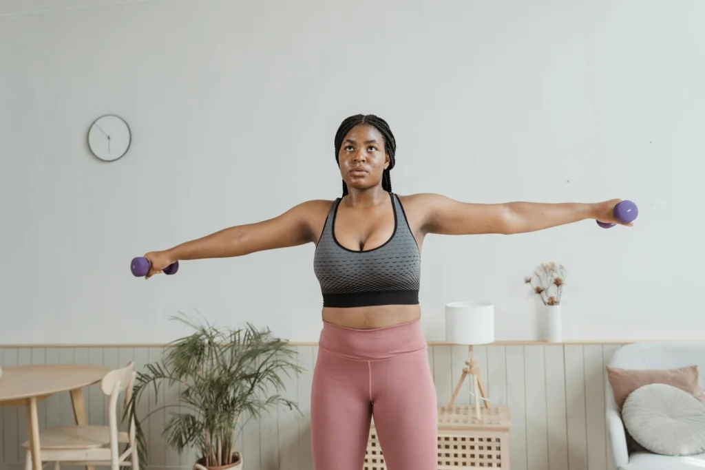 Black woman in activewear performing arm exercises indoors with dumbbells.