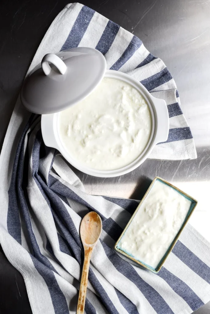 Top view of homemade yogurt in a ceramic pot, styled with a wooden spoon and striped cloth.