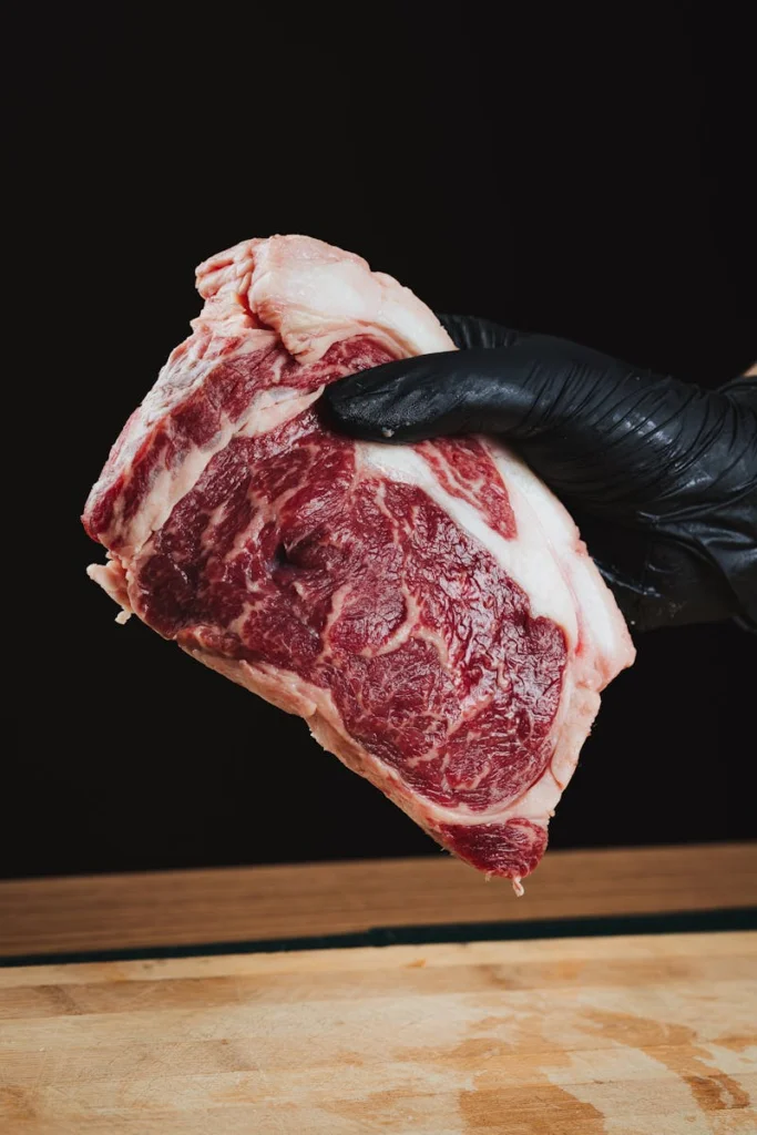 Close-up of a raw ribeye steak held by a gloved hand on a wooden surface.