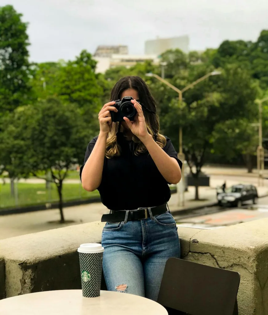 Woman in black shirt taking a photo outdoors with DSLR camera, coffee cup on table.