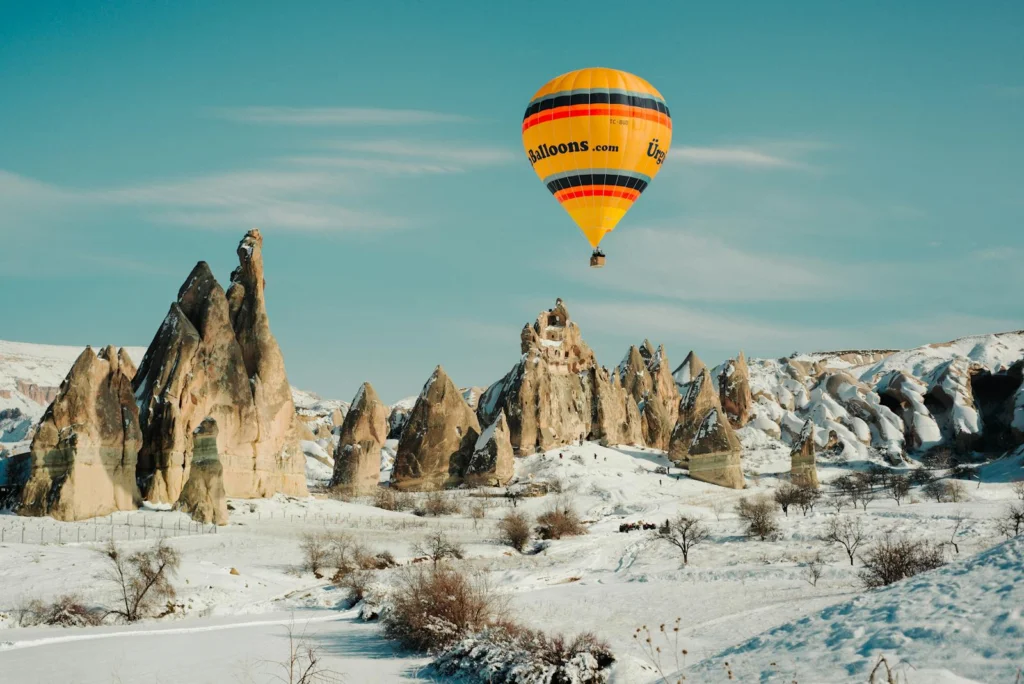 Explore the mesmerizing snow-covered landscapes and unique rock formations of Cappadocia with a vibrant hot air balloon floating overhead.