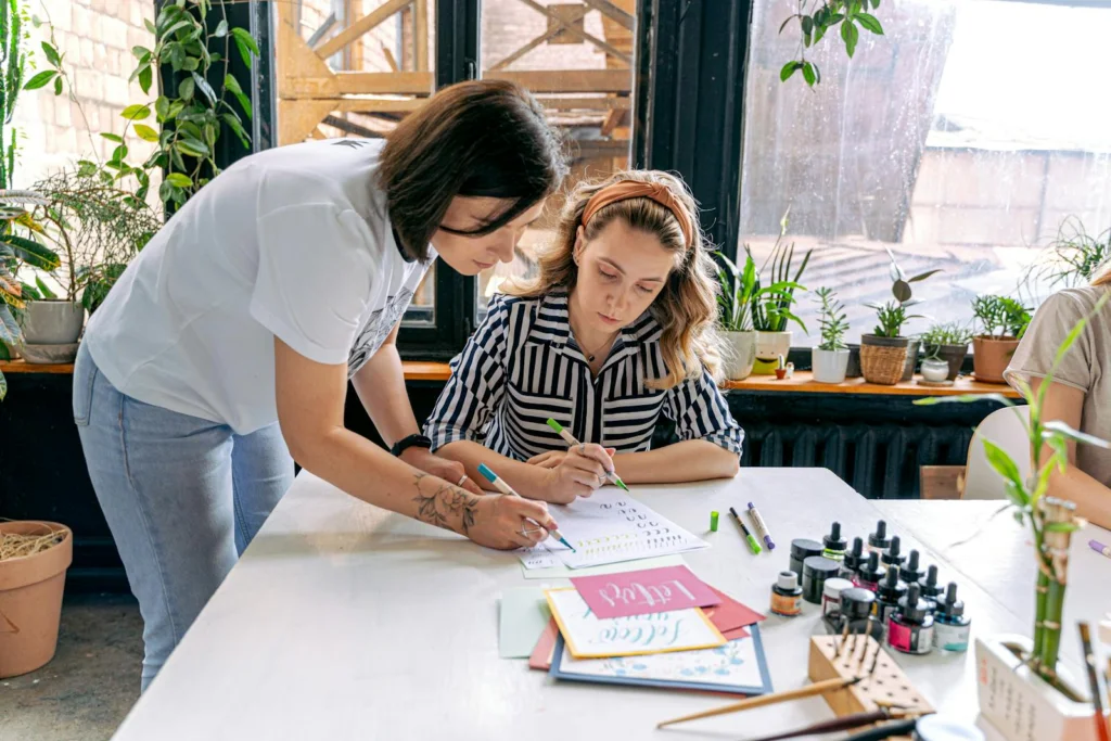 Instructor guiding student in an art workshop. Creative atmosphere with plants and art supplies.