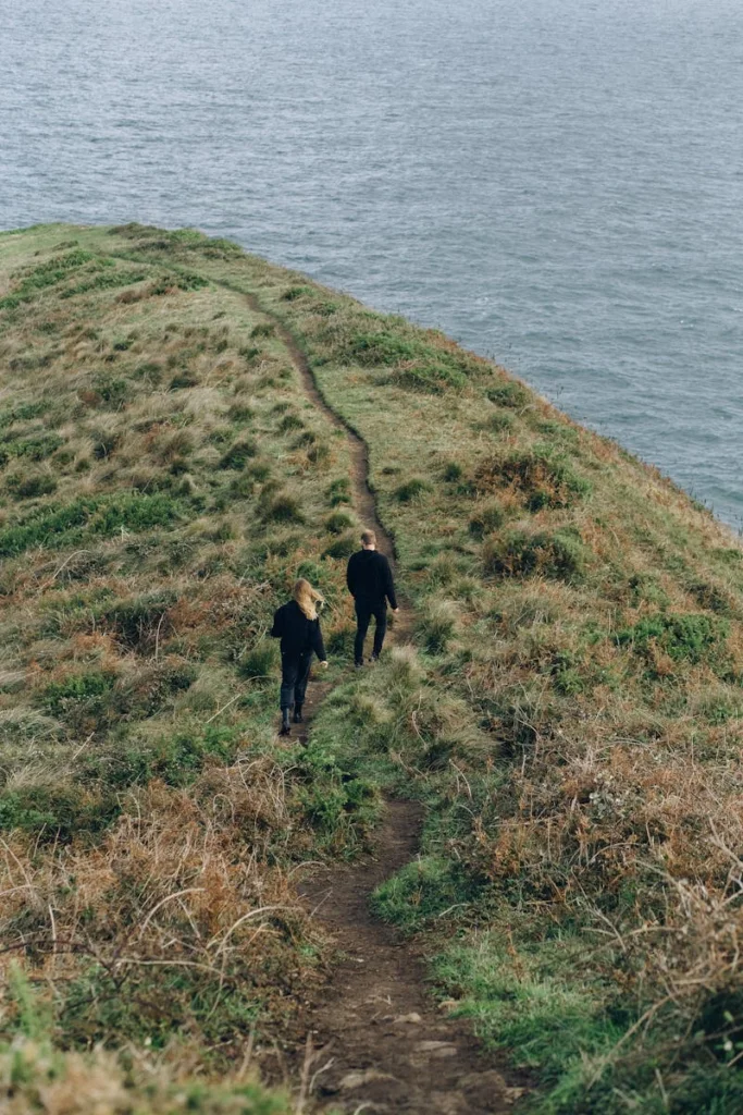 Two hikers walking on a scenic coastal cliff trail beside the ocean under a cloudy sky.