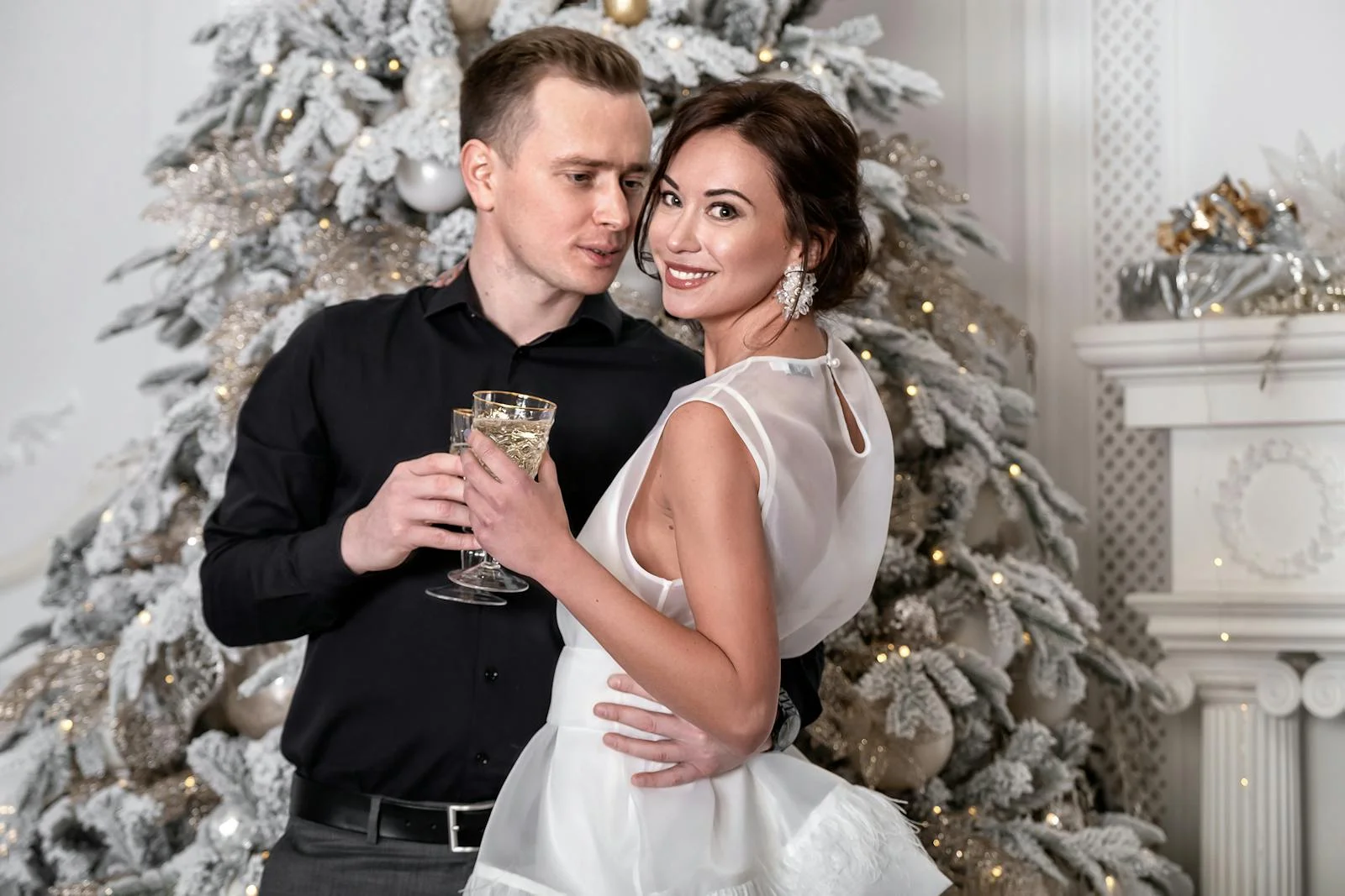 A couple elegantly dressed celebrate Christmas by a festive tree indoors, sharing a joyful moment.