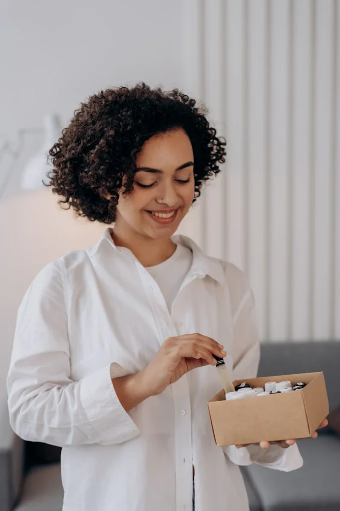 Smiling woman holding essential oil bottles, creating a calming ambiance indoors.