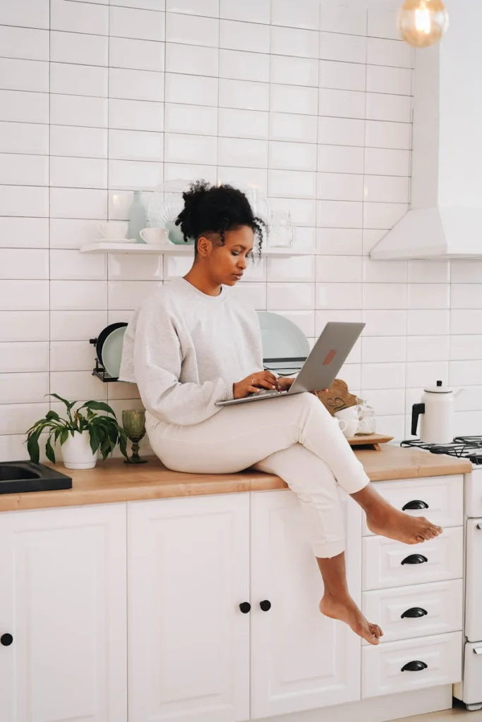African American woman sitting on kitchen counter and typing on a laptop.