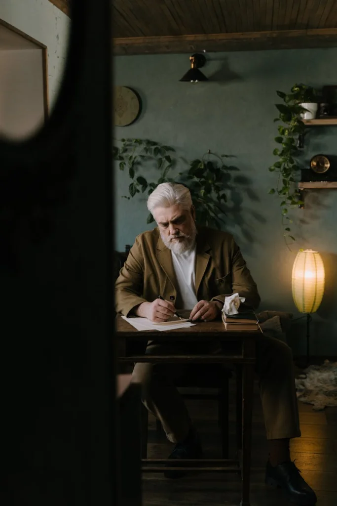 Elderly man with white hair and beard writing at a wooden desk in a warmly lit room with plants.