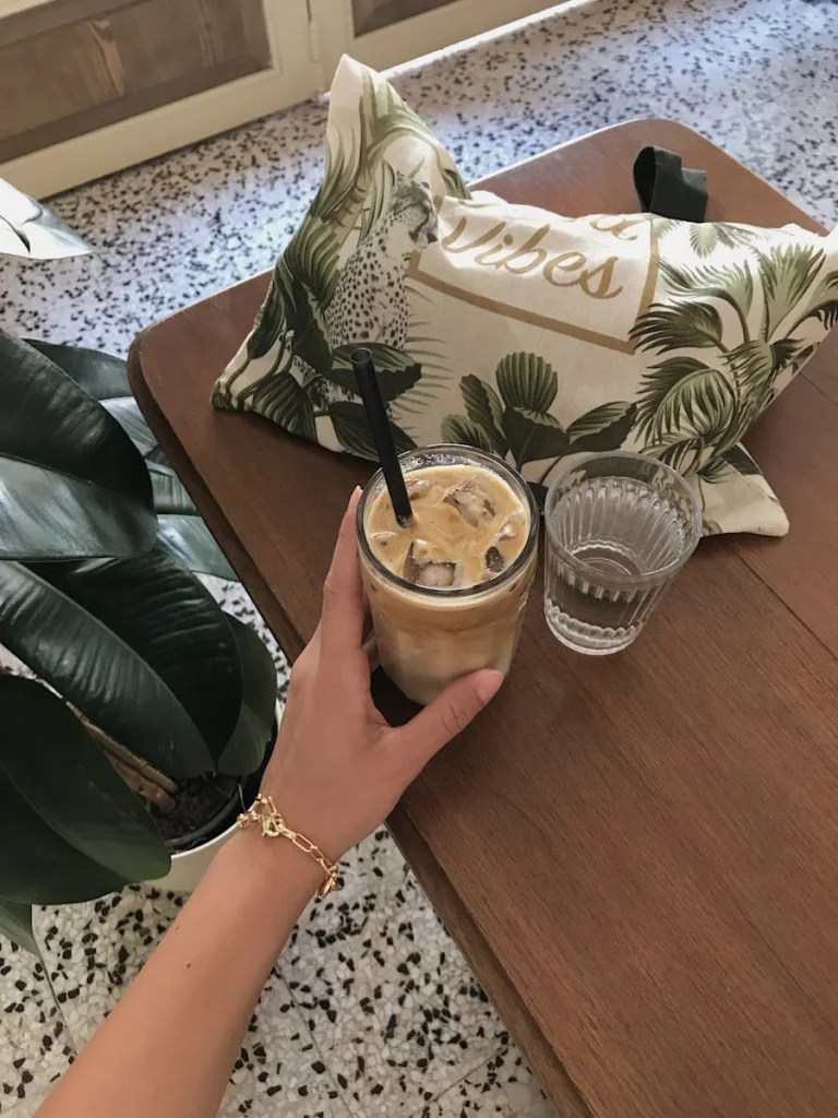 A person's hand holding an iced coffee with a straw placed on a wooden table indoors, featuring clear glass and greenery.