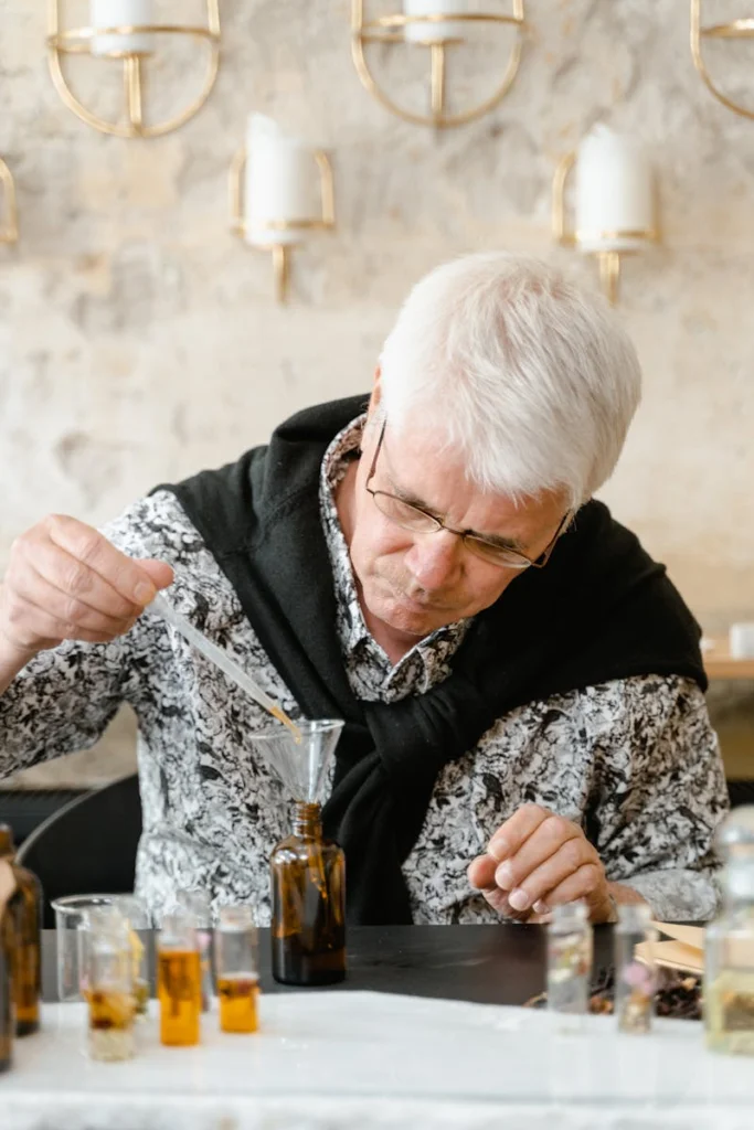 Elderly man carefully crafting a perfume blend in an indoor workspace with various bottles.