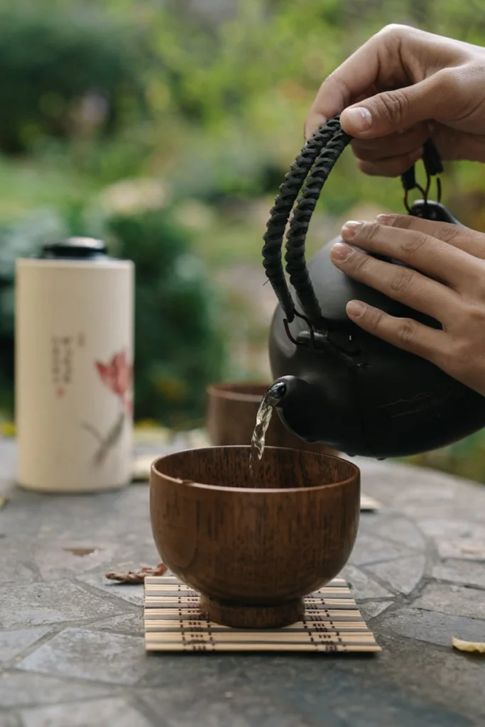 Hands pouring hot tea from a teapot into a wooden cup on a bamboo mat, outdoors.