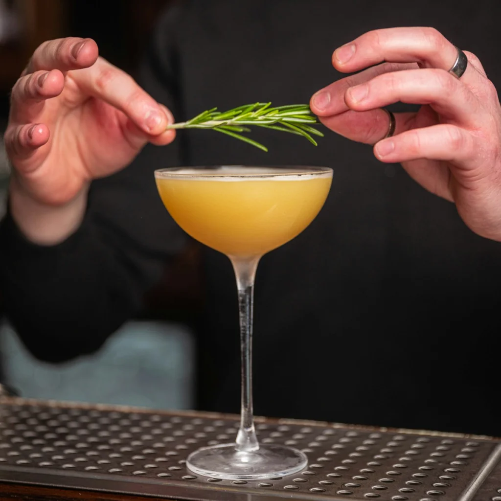 a man is holding a rosemary garnish in a cocktail glass