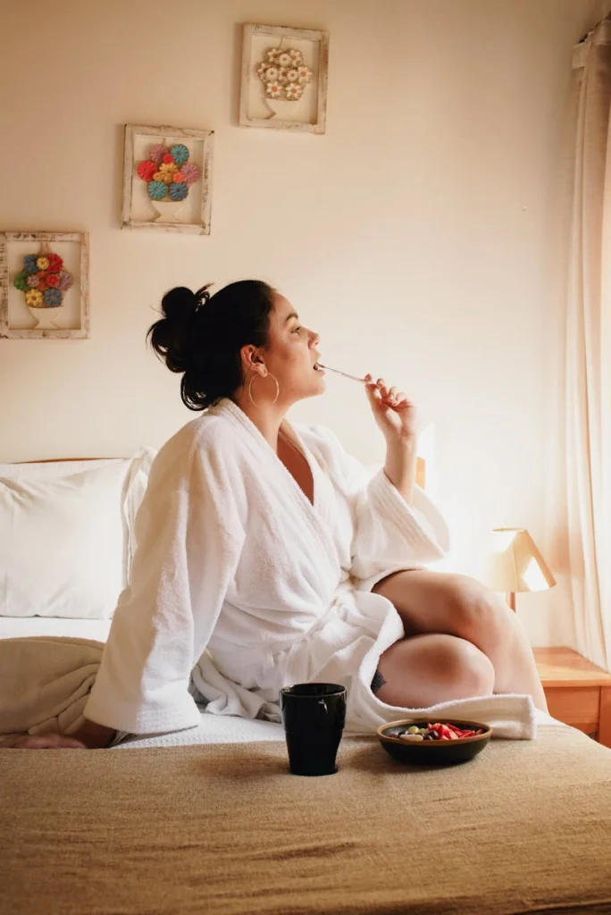 Woman in white robe enjoying breakfast in a cozy bedroom with natural light.