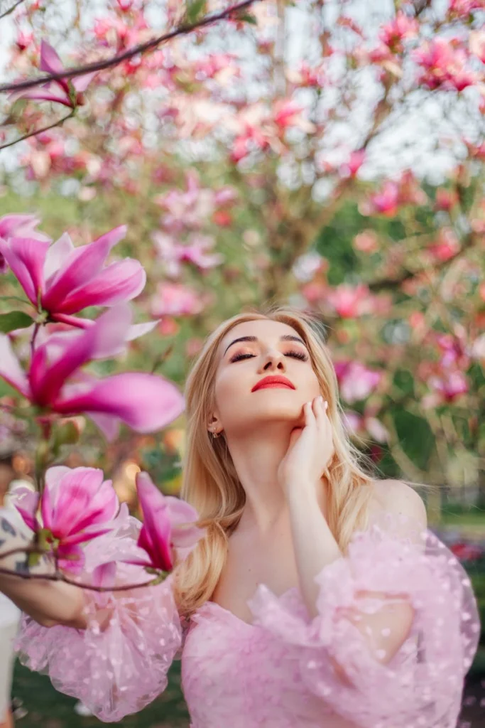 A woman in a pink off-the-shoulder dress poses elegantly amidst blooming magnolia trees outdoors.