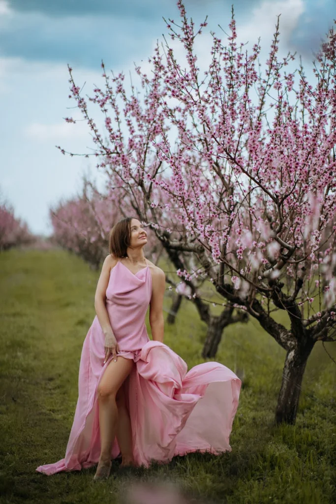 A woman in a flowing peach dress posing amidst blooming peach trees in springtime.