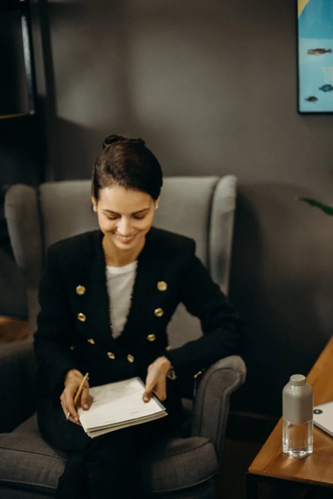 Smiling woman writing notes at home in chair, holding notebook and pen.