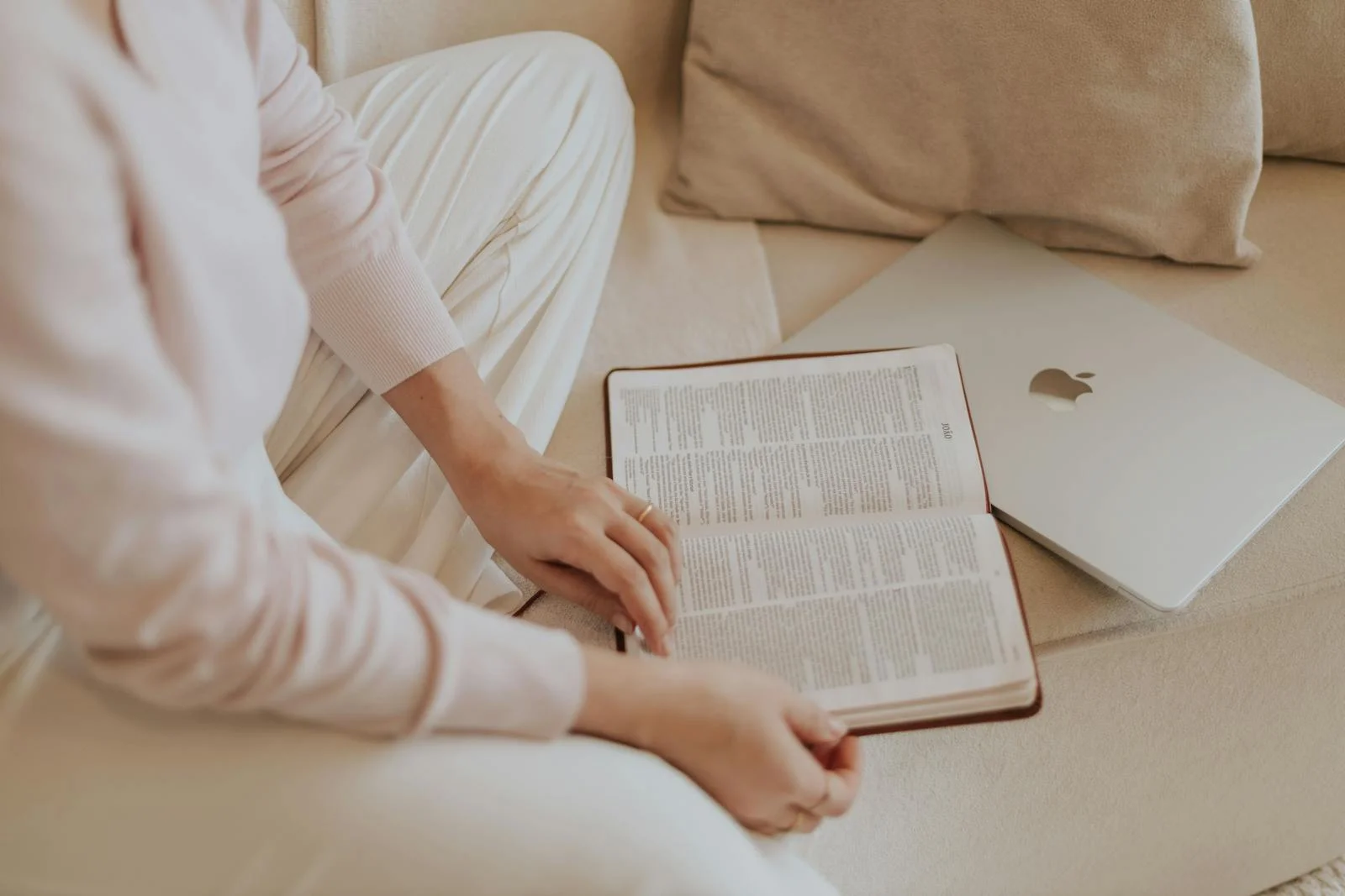 A woman in casual attire reads a book beside a laptop while sitting on a cozy sofa.