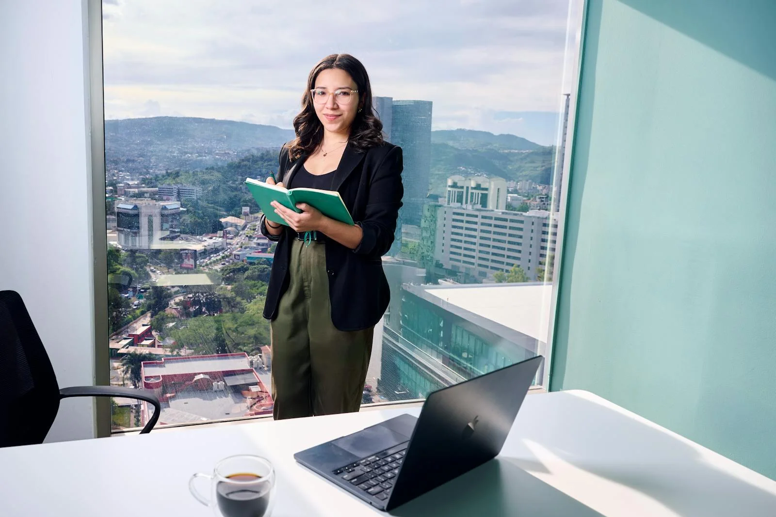 Businesswoman in office holding notebook by window overlooking cityscape.
