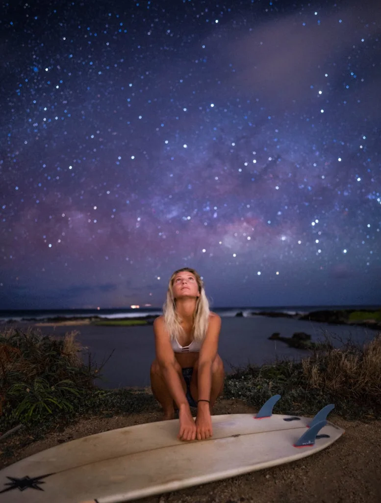 A woman with a surfboard gazes at a starry night sky on a beach, capturing an outdoor evening adventure.