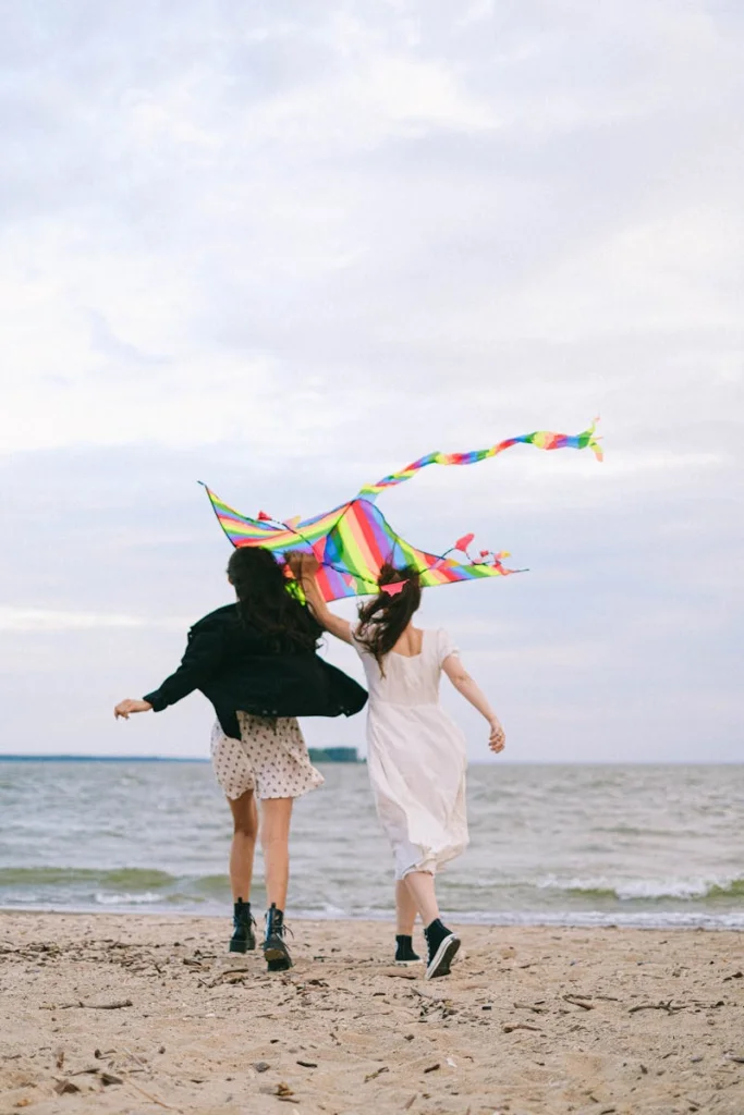 Two women joyfully running with a rainbow kite on a sandy beach, embodying freedom and playfulness.