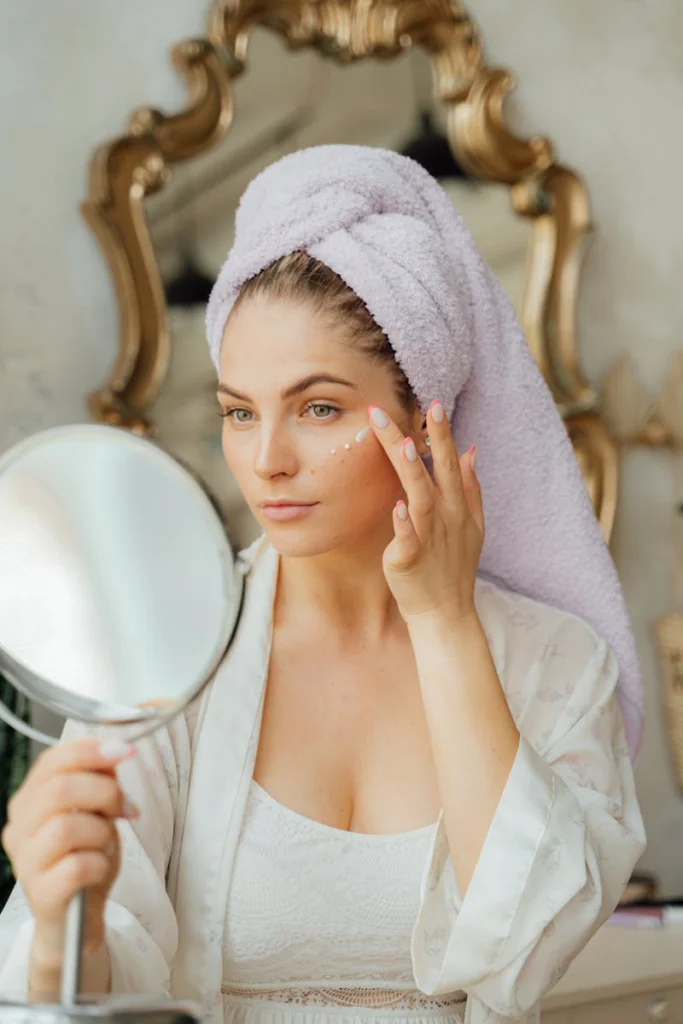 A woman in a towel turban applies face cream while holding a mirror indoors.