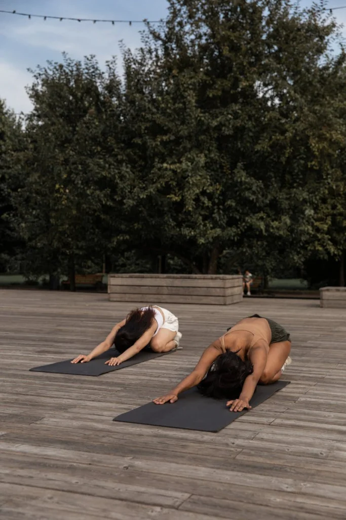 Two women practicing yoga in a serene park setting, outdoors on wooden boards.