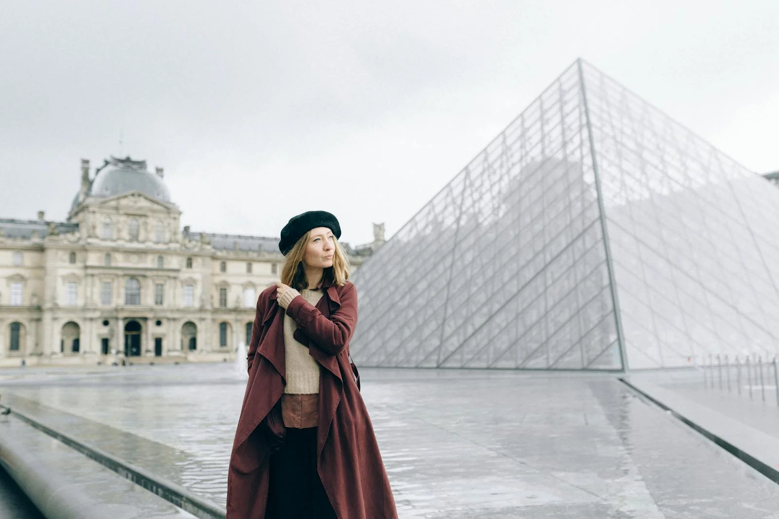 A stylish woman stands near the iconic Louvre Pyramid in Paris, France, on a cloudy day.