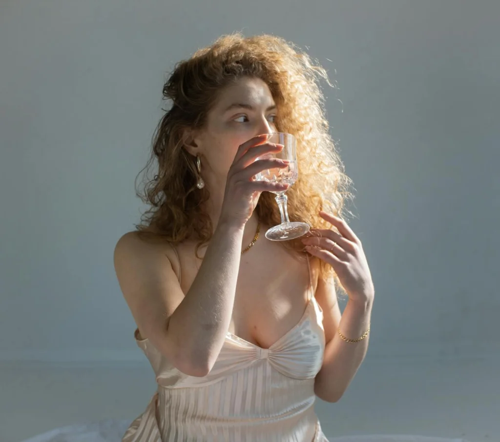 Young female with curly hair bare shoulders and chest looking away while drinking water on white background