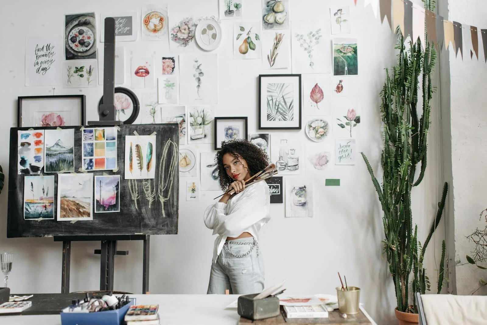 A young female artist poses with paintbrushes in her creative studio filled with watercolor artworks.