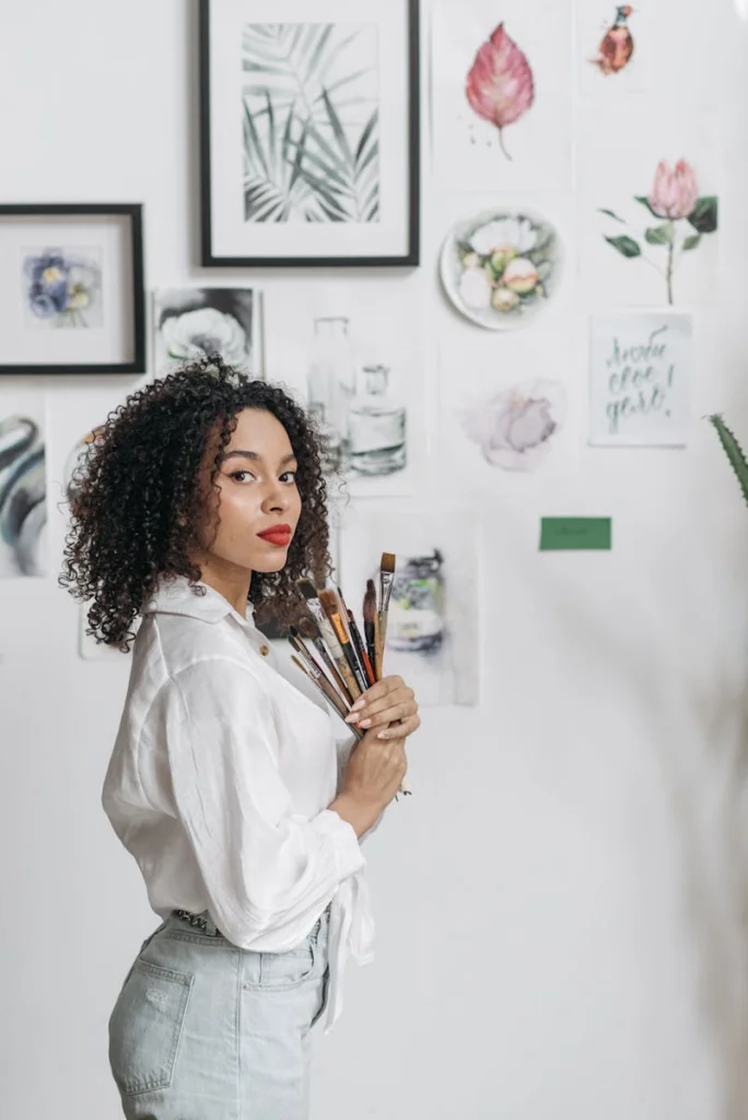 Elegant woman artist holding paintbrushes, surrounded by watercolor paintings indoors.